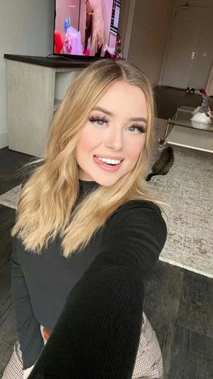 Blonde woman in black long-sleeve top takes selfie in living room with cat in background.