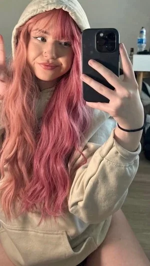 Woman with pink hair in beige hoodie taking mirror selfie indoors.