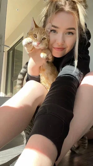 Woman with blonde hair holding a cat, wearing black long sleeves and shorts, sitting outdoors.