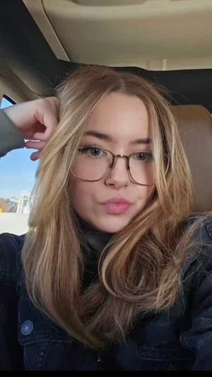 Woman with blonde hair and glasses, wearing a dark jacket, sitting in a car.