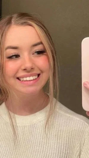 Woman with blonde hair in a white knit sweater, smiling in a mirror selfie indoors.