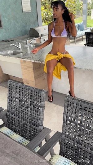 Woman in purple bikini top and yellow wrap posing outdoors on patio near kitchen island.