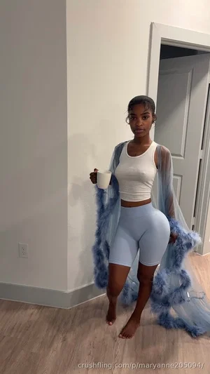 Woman in white crop top and light blue shorts, holding mug, standing indoors with blue feathered