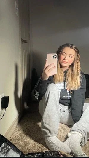 Woman with blonde hair in a gray hoodie and sweatpants sitting on floor, taking mirror selfie.