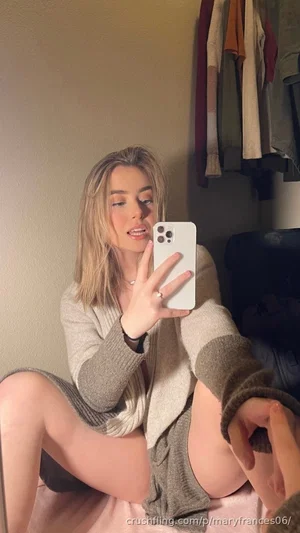 Woman with blonde hair in a gray cardigan and pink skirt, sitting indoors taking a mirror selfie.