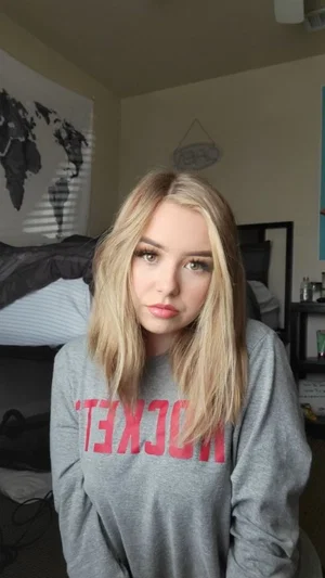 Blonde woman in gray sweatshirt with red text, sitting in bedroom.