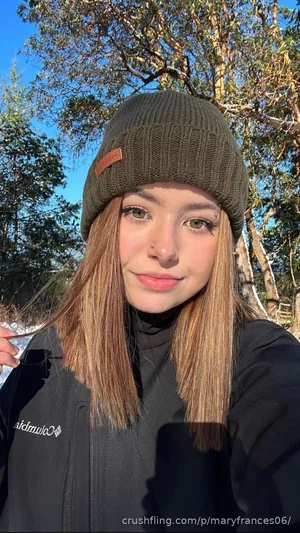 Woman with long hair wearing a beanie and black jacket outdoors in a wooded area.