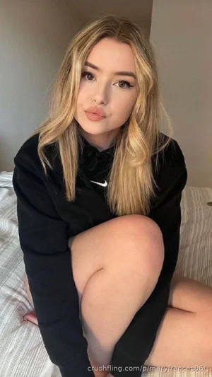 Woman with blonde hair in black hoodie sitting on bed, legs crossed.