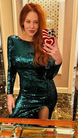 Woman with red hair in a teal sequin dress takes mirror selfie in a room with patterned walls.