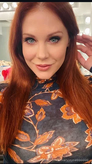 Woman with red hair and blue eyes wearing a floral high-neck top, indoors near a mirror with lights.
