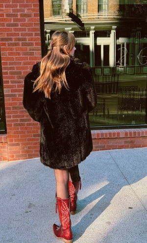 Woman in black fur coat and red boots on city sidewalk.