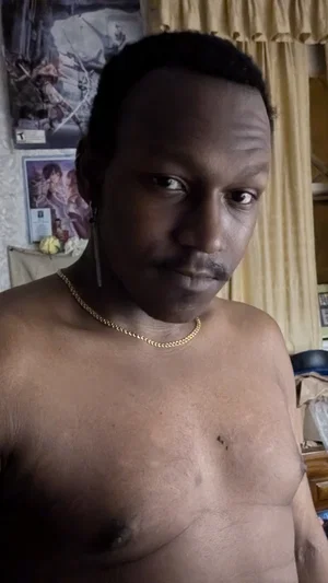 Shirtless man with gold chain, standing indoors near curtains and posters.