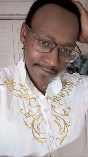 Man with glasses and mustache wearing white shirt with gold embroidery, indoors.