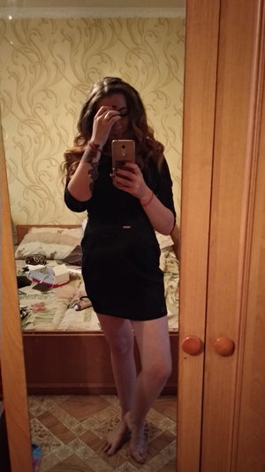 Woman in short black dress taking mirror selfie in bedroom.