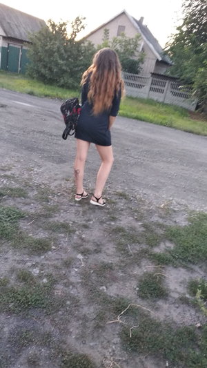 Woman with long hair in black dress standing outdoors on gravel, facing away.