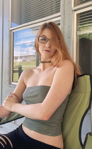 Woman with red hair and glasses in a strapless top, sitting outdoors near a window.