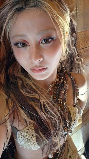 Woman with wavy blonde hair wearing crochet top and beaded necklace indoors.