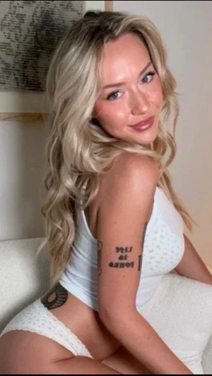 Blonde woman in white lingerie sitting on a bed, showing tattoos on arm and side.