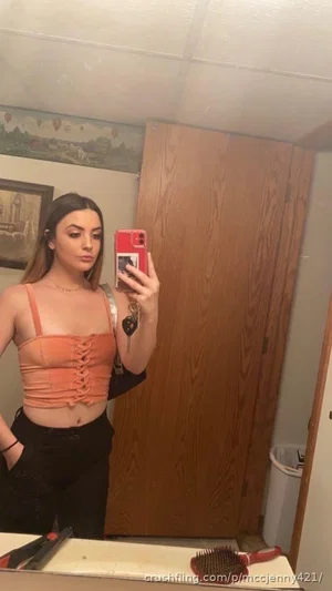 Woman in orange lace-up top and black pants takes mirror selfie in indoor room.