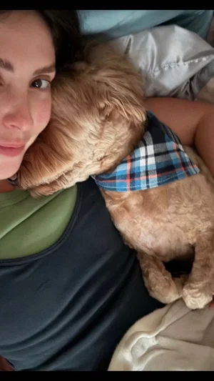 Woman in dark tank top lying in bed with small dog wearing plaid bandana.