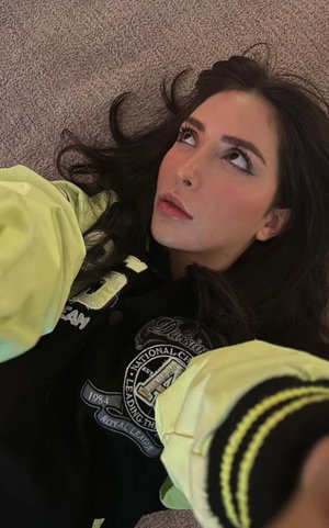 Dark-haired woman in varsity jacket taking overhead selfie indoors.