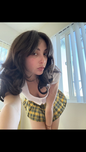 Woman with dark hair in a yellow plaid skirt and white top, posing indoors near a window.