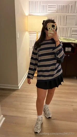 Woman in striped sweater and black skirt takes mirror selfie in bedroom.
