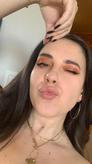 Woman with long dark hair and gold jewelry posing for a close-up selfie indoors.