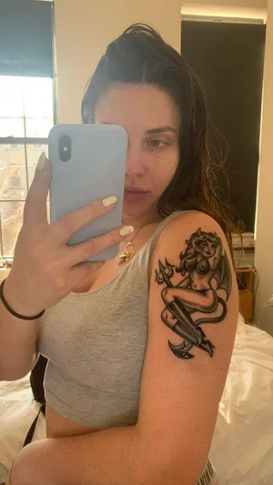 Woman with tattoo on arm wearing gray tank top, taking mirror selfie in bedroom.
