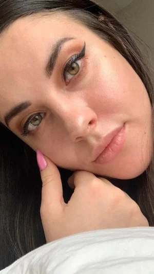 Woman with dark hair and green eyes resting face on hand, wearing white top, indoors.