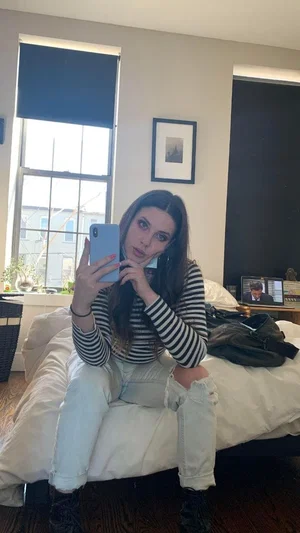 Woman with long dark hair in striped shirt and ripped jeans sits on bed taking mirror selfie in