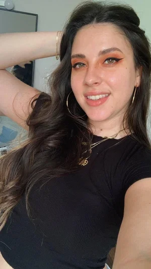Woman with long dark hair and makeup wearing black top, smiling in indoor setting.