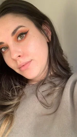 Woman with dark hair and makeup wearing a gray top, close-up selfie indoors.