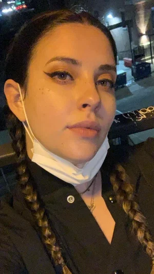 Woman with braided hair and dark makeup, wearing black shirt and face mask under chin, outdoors at