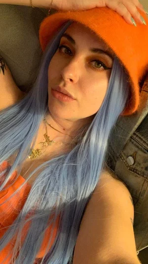 Woman with blue hair and orange hat, wearing orange top, indoors.
