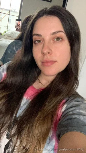 Woman with long dark hair in tie-dye shirt taking selfie in bedroom.