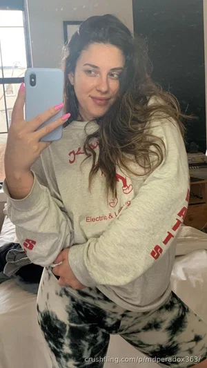 Woman with long brown hair in a gray sweatshirt and tie-dye pants taking a mirror selfie in a