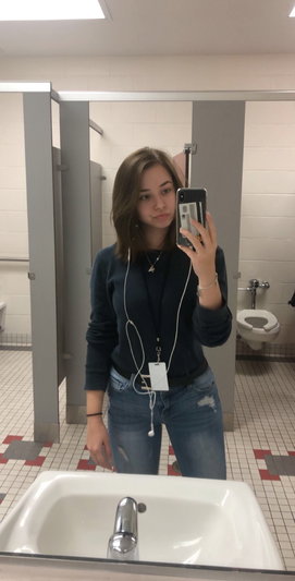 Woman in sweater and jeans taking mirror selfie in bathroom.