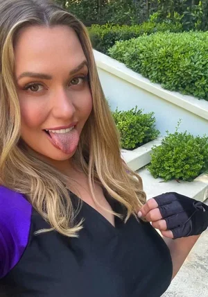 Woman with blonde hair in black and purple top, sticking tongue out, outdoors with greenery.