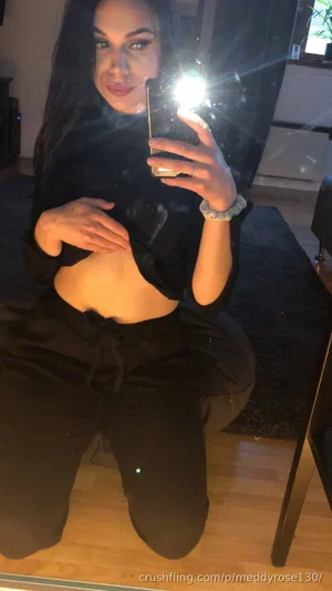 Woman with long dark hair in black crop top and pants, kneeling in mirror selfie indoors.