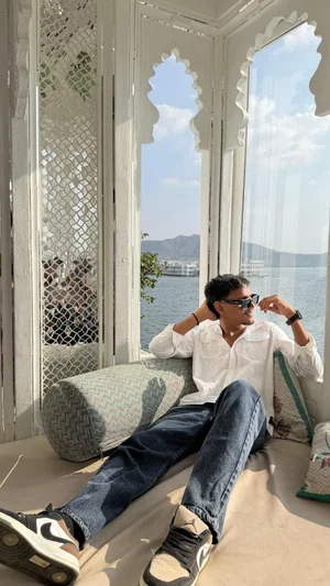 Man in white shirt and jeans lounging on cushioned seat by ornate window with water view.