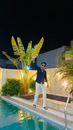 Man in dark shirt and white pants posing by pool at night with plants and lights.