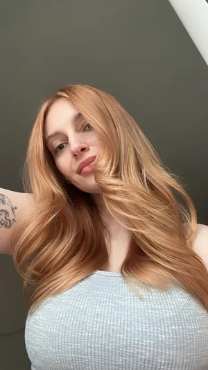 Woman with long blonde hair wearing a light blue top, taking a selfie indoors.