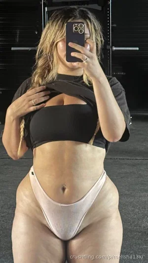 Woman in black crop top and light pink thong taking mirror selfie in gym.