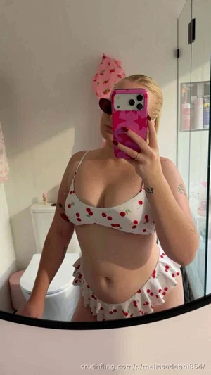 Woman in cherry-print bikini taking mirror selfie in bathroom.