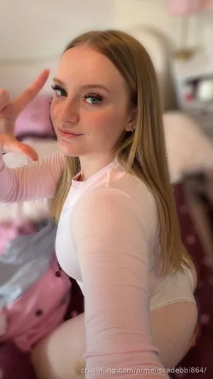 Woman with blonde hair in pink long-sleeve top, posing in bedroom with hand gesture.