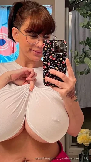 Woman with glasses and ponytail in white top, holding phone for mirror selfie in bedroom.