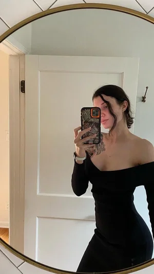 Woman in black off-shoulder dress taking mirror selfie in indoor setting.