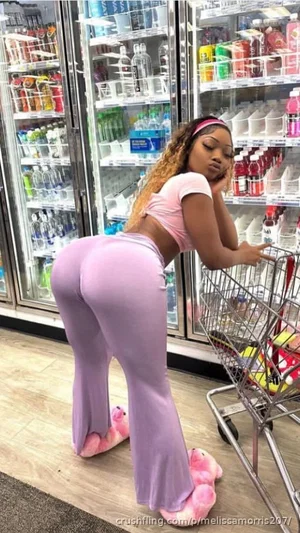 Woman in pink crop top and purple pants posing with shopping cart in grocery store.