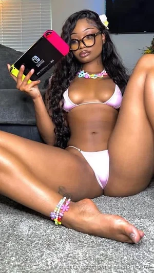 Woman in pink bikini posing on carpet, holding phone, long curly hair, glasses.